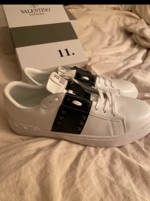 Valentino Rockstud vita sneakers - Säljer ett par Valentino Garavani Rockstud sneakers i vitt skinn med svart detalj och ikoniska nitar på sidan. Riktigt snygga och stilrena sneakers för dig som gillar exklusiva detaljer. De köptes på Plick och är därför osäker på om de är äkta! De är i storlek 43 och aldrig använda❤️boxen på sista bilden medföljer.