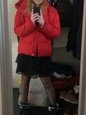 Röd pufferjacka med huva - Säljer en röd pufferjacka med huva och dragkedja framtill. Jackan har en boxig passform och är vadderad för extra värme. Perfekt för kyliga dagar och enkel att styla med både jeans och kjol. Snygga snören i huvan för justering.