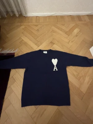Stickad tröja från Ami Paris - Säljer en mörkblå stickad tröja från Ami Paris med deras ikoniska vita hjärta och A på bröstet. Tröjan har rund halsringning och långa ärmar. Perfekt för dig som gillar stilrena plagg med en twist.