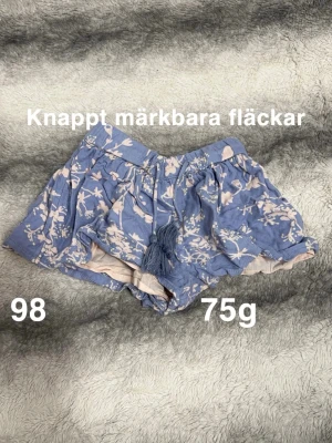 Blommiga blå shorts med snörning - Säljer ett par blå shorts med blommigt mönster och dekorativ snörning med tofsar framtill. De har en lös passform och är perfekta för varma dagar. Materialet känns mjukt och luftigt, vilket gör dem riktigt sköna att ha på sig.🌸 Knappt märkbara fläckar. Storlek 98