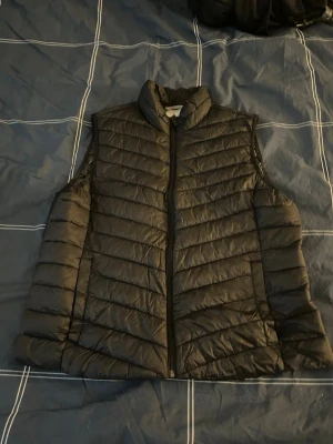 Svart dunväst från McKinley - Säljer en svart dunväst från McKinley i storlek 46. Västen har quiltad design, hög krage och dragkedja framtill. Perfekt att slänga över en hoodie eller tröja när det är kyligt ute. Vet ej om det är dam eller herr modell.