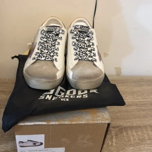 Golden Goose sneakers med glitterstjärna - Snygga Golden Goose sneakers i vitt skinn med grå mockadetaljer på tån, rosa glittrig stjärna på sidan och metallicrosa häl. Skorna har svarta och vita snören med leopardmönster och platt sula. Perfekt för dig som vill sticka ut med coola detaljer.