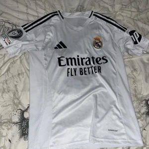 Real Madrid Bellingham 5 matchtröja - Real Madrid fotbollströja i vitt med svarta detaljer, Adidas-logga och klubbmärke på bröstet. Nummer 5 och Bellingham på ryggen. Champions League-märke på ärmen och 'Emirates Fly Better' tryck på framsidan. Tillverkad i lätt och ventilerande material.