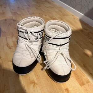 Vita Moon Boots med snörning - Moon Boots i vit färg med svarta detaljer och snörning framtill. De har en rund tå och platt sula, samt en vadderad och quiltad ovandel i syntetmaterial. Perfekta för kalla vinterdagar och har en ikonisk retrostil.