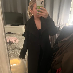 Svart lång kappa från Ellos - Säljer en svart, stilren och lång kappa från Ellos i storlek 34. Jackan har bred krage och knäppning framtill, med en snygg loose passform som ger en chill vibe. Perfekt för dig som gillar minimalistisk stil och vill ha något som funkar hela säsongen. Endast använd några gånger.