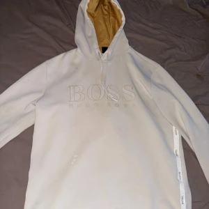 Vit hoodie från Hugo Boss med logga - Snygg vit hoodie från Hugo Boss med stor broderad logga på bröstet och kontrastfärgad beige insida på huvan. Tröjan har långa ärmar, snörning i huvan och dekorativa band med logga längs sidan. Perfekt för en chill och trendig look.