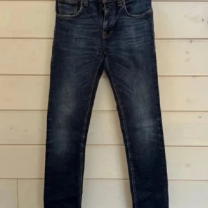 Nudie Jeans blå denim 31/34 - Snygga blå jeans från Nudie Jeans med klassisk femficksdesign och orangea sömmar. Jeansen har en rak passform och subtila slitningar framtill samt Nudie-logga på bakfickorna. Perfekta för dig som gillar stilrena och tidlösa jeans.