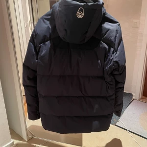 Mörkblå Sail Racing pufferjacka - Säljer en mörkblå pufferjacka från Sail Racing med huva och dragkedja framtill. Jackan har två bröstfickor med dragkedja och logotyp på både bröstet och ärmen. Perfekt för kalla dagar med sin tjocka och vadderade design.