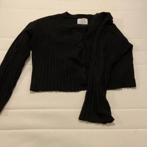 Svart ribbad kofta från Zara - Svart croppad kofta från Zara med ribbad struktur och stora knappar framtill. Koftan har långärmade ärmar och är tillverkad i ett stickat material som känns mjukt och skönt. Perfekt för dig som gillar en enkel men snygg look.