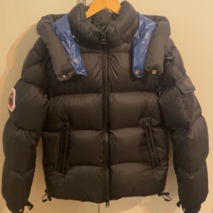 Svart pufferjacka från JACK1T, S - Supersnygg svart pufferjacka från JACK1T med blå luva inuti och loggan stort på ärmen. Jackan har dragkedja framtill, två fickor med dragkedja och justerbara snoddar nedtill & vid luvan. Håller värme väldigt bra och snygg till vintern. Skriv om ni vill ha fler bilder på osv!