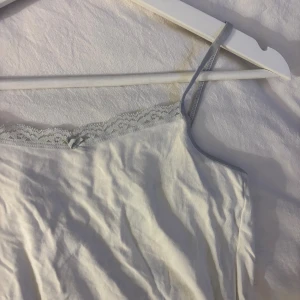 Vit linnetopp med spetskant - Säljer en vit linnetopp med tunna axelband och ljusgrå spetskant upptill. Toppen har en liten rosett framtill och är gjord i mjuk bomull. Perfekt för sommarens varma dagar eller som layering-piece. Enkel och stilren design med feminin touch.