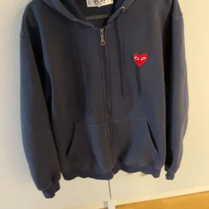 Mörkblå hoodie Comme des Garçons Play - Mörkblå hoodie från Comme des Garçons Play med dragkedja och huva. Ikoniskt rött hjärta med ögon broderat på bröstet. Mjuk bomullskvalitet och känguruficka framtill. Perfekt för en chill och stilren look.