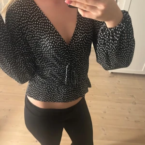 Svart prickig blus med snörning - Säljer en svart blus med vita prickar och v-ringad hals. Blusen har långa puffiga ärmar med resår vid handlederna. Materialet är lätt och luftigt, sparsamt använd.