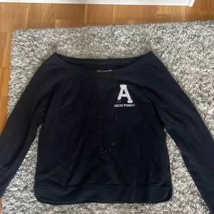 Svart sweatshirt från Abercrombie & Fitch - Svart sweatshirt från Abercrombie & Fitch med stort vitt A och broderad logga på bröstet. Tröjan har bred halsringning och långa ärmar vilket gör den liten of sholder, perfekt för en chill och avslappnad look. Mjuk insida och klassisk passform.