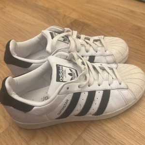 Adidas Superstar vita sneakers - Klassiska Adidas Superstar sneakers i vitt med svarta ränder och svart häl. Skorna har snörning, rund tå och platt sula. Tillverkade i skinn med ikonisk skalformad tå. Perfekta för dig som gillar retrostil och streetwear.
