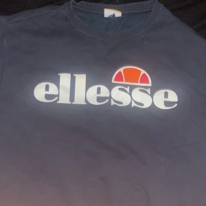 Mörkblå sweatshirt från Ellesse - Säljer en mörkblå sweatshirt från Ellesse med stort vitt logotryck och orange detalj framtill. Tröjan har rund halsringning och är i mjukt material, perfekt för en chill och sportig look.