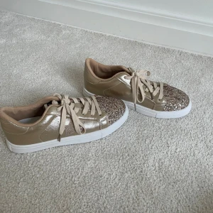 Guld glittriga sneakers med stjärndetalj - Snygga guld sneakers med glittriga detaljer på tån och en glittrig stjärna på sidan. Skorna har snörning, vit platt sula och skimrande syntetmaterial som ger en riktigt trendig look. Perfekta för dig som vill sticka ut lite extra.