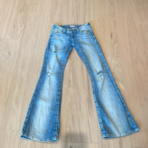 Blå bootcut jeans Gina Tricot XS - Bootcut jeans från Gina Tricot i ljusblå tvätt med slitna detaljer och snygga broderade bakfickor. Klassisk femficksmodell med låg midja och dragkedja. Perfekt för dig som gillar retrovibbar och avslappnad stil.