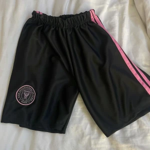 Inter Miami svarta shorts med rosa detaljer - Svarta sportiga shorts från Inter Miami med rosa broderad klubbmärke på benet och rosa ränder längs sidorna. Elastisk midja för skön passform. Tillverkade i 100% polyester, perfekta för fotboll eller chill. Snygg och modern look med klubbens logga.