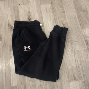 Svarta mjukisbyxor från Under Armour - Svarta mjukisbyxor från Under Armour med broderad logga på benet. Byxorna har resår och snörning i midjan samt muddar vid bensluten. Tillverkade i mjukt material som är perfekt för chill eller träning. Loose fit för extra komfort.