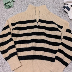 Randig stickad half zip-tröja beige/svart - Beige och svart randig stickad tröja med half zip och hög krage. Tröjan har långa ärmar och breda muddar vid ärmslut och nederkant. Perfekt för lager-på-lager och chill höst- eller vinterstil. Bra skick bara lite noppig☺️