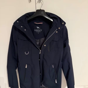Mörkblå vindjacka från Snoot Stockholm - Snygg mörkblå vindjacka från Snoot Stockholm med flera fickor, dragkedjor och coola metalldetaljer. Jackan har huva som går att ta av, hög krage och smarta funktioner som D-ring och justerbara ärmslut. Perfekt för dig som gillar stil och funktion.