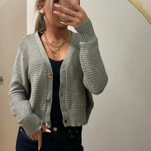 Grå ribbad kofta med knappar - Snygg grå kofta med ribbad struktur och tre stora knappar framtill. Modellen är croppad med breda muddar vid ärmslut och nederkant, samt v-ringad hals. Perfekt att styla över ett linne eller t-shirt för en chill look.