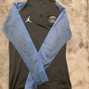 PSG träningströja - Svart träningströja från PSG x Jordan med blå långärmade ärmar och diskret mönster. Halv dragkedja framtill, PSG-logga och Jumpman på bröstet. Materialet är polyester, perfekt för fotbollsträning. Röd, vit och blå detalj vid nacken. ( Frankrike ) sista pris 350 kr