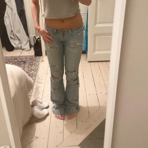 Ljusblå slitna bootcut jeans - Säljer ett par ljusblå lee jeans med bootcut och låg midja. Jeansen har flera slitningar och hål på både lår och knän för en riktigt edgy vibe. Klassisk femficksmodell och tillverkade i jeansmaterial. Perfekt för dig som gillar en avslappnad och trendig stil. Midja 37 innerben 78