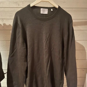 Mörkgrön stickad tröja från Black - En enkel och stilren mörkgrön stickad tröja från Black. Tröjan har rund halsringning och långa ärmar, perfekt för lager-på-lager. Materialet är mjukt och skönt mot huden, och passformen är normal. Perfekt för dig som gillar minimalistisk stil.