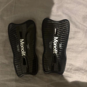 Svarta benskydd Monolit carbon 14 cm - Säljer ett par svarta benskydd från Monolit med cool kolfiberlook. De har en ergonomisk form som sitter skönt runt benet och är tillverkade i lätt och slitstarkt material. Perfekt för dig som vill ha skydd och stil på fotbollsplanen.