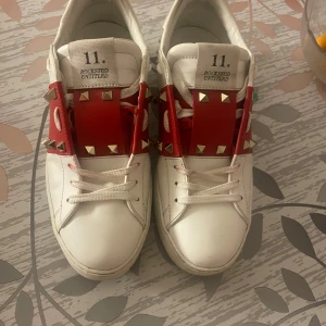 Valentino Rockstud sneakers vit/röd - Snygga Valentino Rockstud sneakers i vitt skinn med röda detaljer och ikoniska nitar över snörningen. Låg modell med platt sula och rund tå. Skorna har coola metalldetaljer och en unik design som sticker ut. Perfekt för dig som gillar statement pieces och exklusiva märken. (Säljs då dom är för små för mig) använda få gånger, pris kan diskuteras!🩷