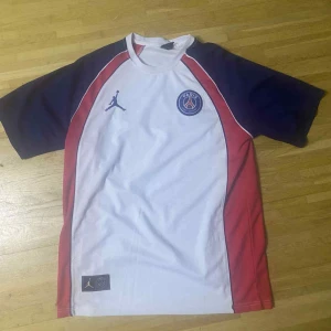 PSG x Jordan vit och marinblå fotbollströja - Snygg PSG fotbollströja i samarbete med Jordan. Tröjan är vit med marinblå ärmar och röda paneler längs sidorna. PSG-logga och Jumpman på bröstet, stor Paris Saint-Germain-tryck på ryggen. Tillverkad i lätt och ventilerande material.