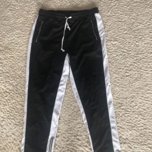 Svarta täckbyxor med vita ränder - Svarta täckbyxor med breda vita ränder längs sidorna, snörning i midjan och dragkedjor både vid fickorna och vid bensluten. Byxorna har en sportig look och är gjorda i ett glansigt syntetmaterial som känns lätt och smidigt.