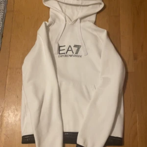 Vit hoodie från Emporio Armani EA7 - Snygg vit hoodie från Emporio Armani EA7 med svart logga på bröstet och svarta detaljer med logotyp på ärmslut och nederkant. Har tyvärr inte använt den så mycket så det är därför jag säljer den nu!