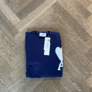 Mörkblå stickad tröja från Ami - Snygg mörkblå stickad tröja från Ami med ett vitt hjärta och bokstaven A på bröstet. Tröjan har rund hals, långa ärmar och ribbade muddar vid ärmslut och nederkant. Perfekt för dig som gillar stilrena detaljer och vill ha något som sticker ut.