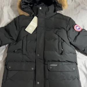 Svart dunjacka från Canada Goose - Helt ny oanvänd!!! Säljer en svart dunjacka från Canada Goose med ikonisk patch på ärmen och huva med snygg fuskpäls. Jackan har flera fickor med både dragkedja och knappar, samt en klassisk pufferlook som håller dig varm hela vintern. /stor och lång modell