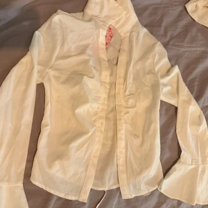 Vit skjorta med snörning från SHEIN - Supersnygg vit skjorta från SHEIN med långa ärmar och breda manschetter. Skjortan har snörning i ryggen och knappar framtill. Materialet är mjukt och lätt, troligtvis polyester.