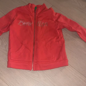 Röd zip-hoodie från Benetton XXS - Snygg röd hoodie från Benetton med glittrigt logotryck på bröstet. Tröjan har dragkedja framtill, två fickor och mjukt bomullsmaterial. Perfekt för dig som gillar färg och vill ha en bekväm och cool look. Sorkel XXS(3-4år)