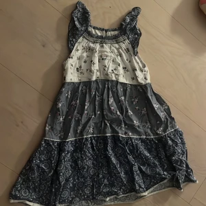 Blommig volangklänning i blått och vitt - Supersöt kortklänning med volanger och breda axelband. Klänningen har flera lager i blått och vitt med småblommigt mönster och smock upptill. Perfekt för sommarens varma dagar. Storlek 122 (6-7år)