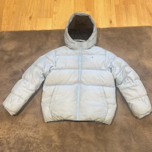 Ljusblå pufferjacka Tommy Hilfiger - Snygg ljusblå pufferjacka från Tommy Hilfiger med huva och dragkedja framtill. Jackan har ett broderat logotypmärke på bröstet och stor Tommy Hilfiger-text på insidan. Tillverkad i polyester och fodrad med ljusblått tyg. Perfekt för kalla dagar.