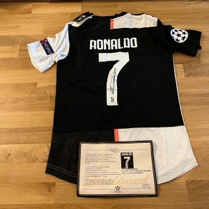 Juventus Ronaldo signerad matchtröja - Juventus fotbollströja med Ronaldo 7, signerad på ryggen. Tröjan är svart och vit med röd detalj, officiella klubbmärken, Champions League- och Respect-patchar på ärmarna samt Jeep-logga framtill. Tillverkad i lätt funktionsmaterial (Climachill).
