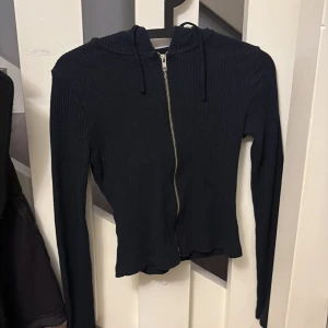 Svart ribbad hoodie från Gina Tricot - Svart ribbad hoodie från Gina Tricot i storlek S. Tröjan har dragkedja framtill, huva med snörning och är långärmad. Perfekt för lager-på-lager och chill dagar. Snygg och enkel att matcha med jeans eller kjol.