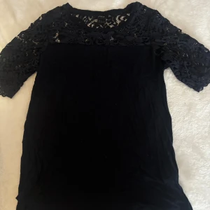 Marinblå blus med spets från Lindex - Snygg svart blus från Lindex med halvtransparent spets upptill och på ärmarna. Blusen har rund halsringning och trekvartsärm med vacker spetsdetalj som ger en cool och feminin vibe. Perfekt att styla med jeans eller kjol.