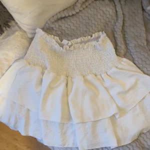 Vit volangkjol med smock i midjan - Supersöt vit kortkjol med bred smock i midjan och dubbla volanger. Kjolen har en luftig känsla och är perfekt för varma dagar. Den stretchiga midjan gör att den sitter bekvämt och volangerna ger en extra feminin touch.