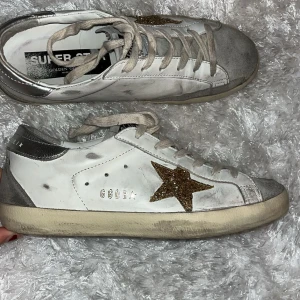Golden Goose skor - Golden Goose sneakers i vitt skinn med grå mockadetaljer och silverfärgad häl. Sidan pryds av en glittrig guldstjärna. Klassisk snörning och platt sula. Perfekta för dig som vill ha en cool och unik look.