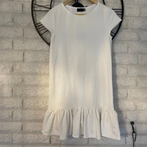 Vit kortklänning med volang från D'Zan - Supersöt vit kortklänning från D'Zan med rund hals och korta ärmar. Klänningen har en clean look och avslutas med en bred volang nedtill som ger extra flärd. Perfekt för dig som gillar minimalistisk stil med en twist.