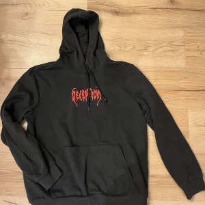 Svart hoodie med röd print Pier One - Svart hoodie från Pier One med stor röd print 'DECEPTION' på bröstet och ryggen, samt grafiskt tryck med läppar och text. Tröjan har huva med snörning, magficka och långa ärmar. Materialet är mjuk bomull, perfekt för en edgy streetstyle-look.