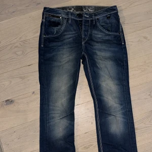 Blå jeans Jack & Jones Rick Curve - Snygga blå jeans från Jack & Jones, modell Rick Curve. Jeansen har en klassisk femficksdesign med kontrastsömmar, knappgylf och coola detaljer som dragkedja på fickan. Passformen är rak och materialet är slitstarkt denim. W31 L32