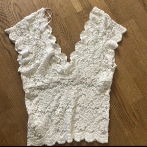 Vit spetsig croppad linnetopp - Säljer en vit croppad linnetopp med v-ringad hals och breda axelband. Toppen är helt täckt av vacker spets med blommigt mönster och har vågiga kanter både upptill och nedtill. 